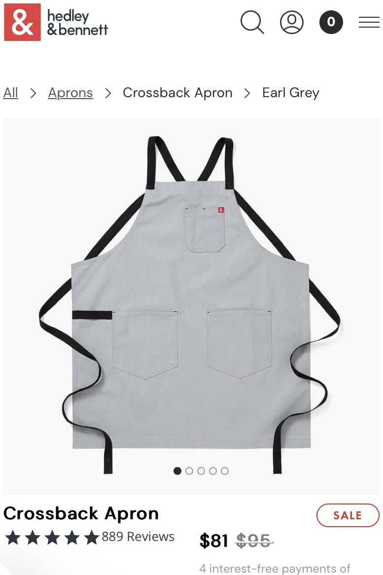 Screenshot from a website of a gray apron with black straps laid flat on a white background. The listing says 'Hedley and Bennett Crossback Apron'